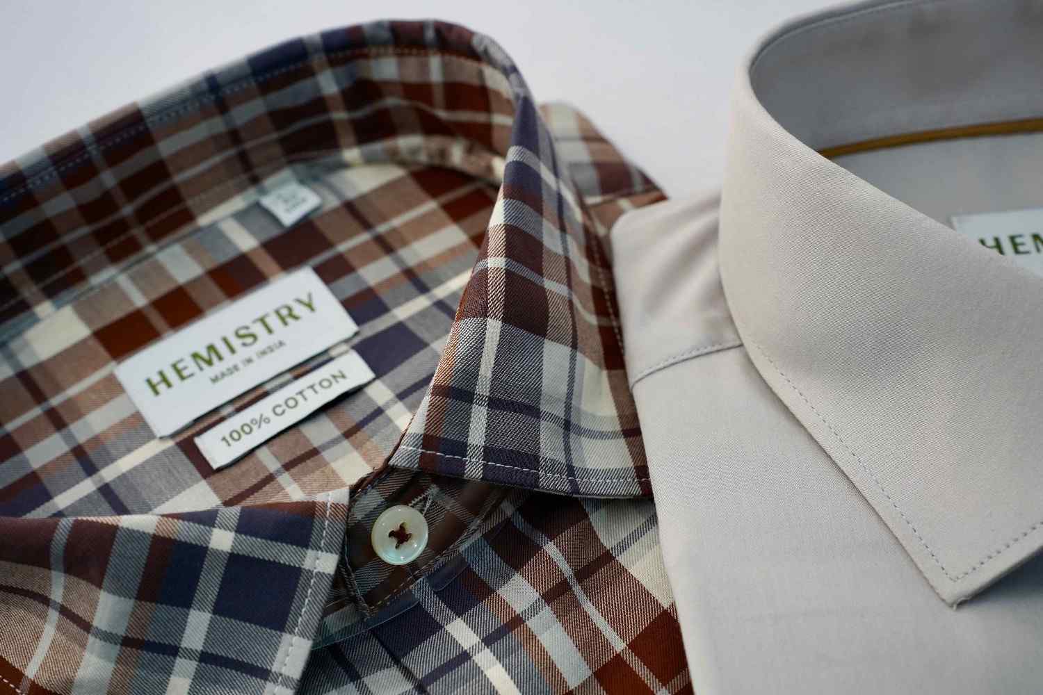 Closeup of two men's shirts, one is brown cream plaid shirt with visible Hemistry Neck label and one grey shirt